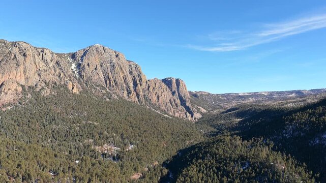 Drone footage glides above dense evergreen forest toward massive rock walls, revealing the Brazos Cliffs towering above a mountain valley with light snow patches and rugged alpine character.