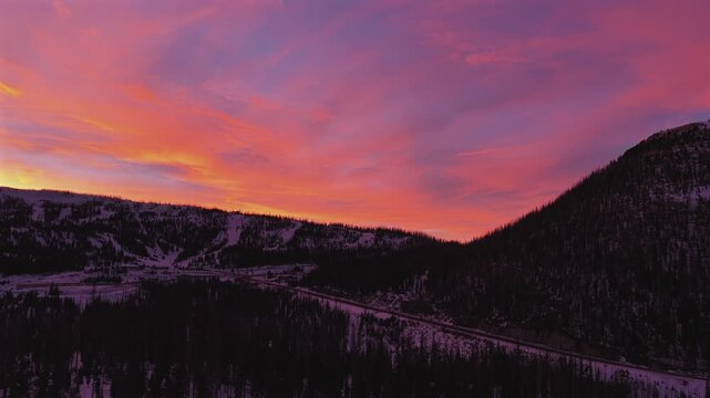 Drone footage of a single car driving along a rural mountain highway as surreal pink and purple sunset colors fill the winter sky, creating a cinematic, isolated mood in the high country.