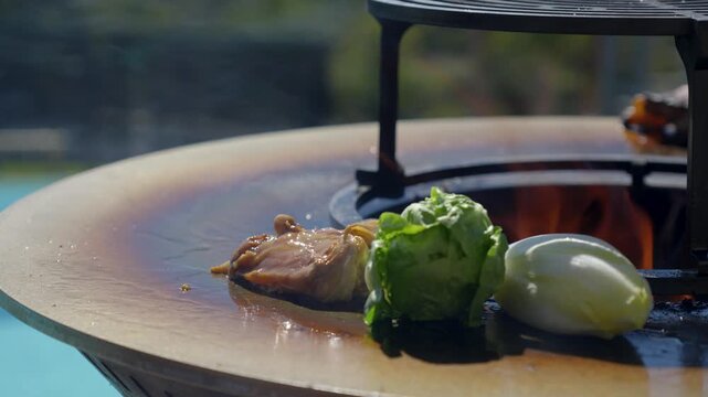 A person uses metal tongs to place cherry tomatoes on the vine onto the hot, circular surface of an outdoor fire pit grill, which already holds vegetables. Focus on gourmet open-fire cooking.