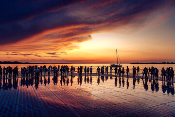 View of a crowd of silhouetted people gathered on a reflective surface, mirroring the fiery sunset sky over the calm sea, Zadar, Zadar County, Croatia.