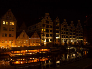 Naklejka premium Tall buildings line the water in Gdansk at night. Lights from windows and a boat create reflections on the surface.