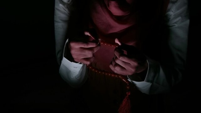 Cinematic overhead shot of a young woman with a headscarf holding prayer beads in a dark atmosphere, expressing spirituality, belief, contemplation, inner strength, and a calm yet emotional mood