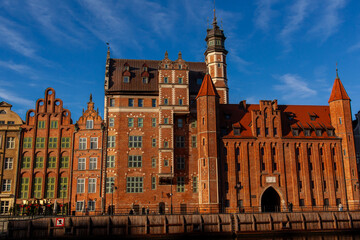 Obraz premium People walk near historic brick buildings by the canal in Gdansk as the sun sets in the evening sky. St. Mary Gate (Brama Mariacka) on the embankment of Motlawa River, Poland. Mariacka Gate