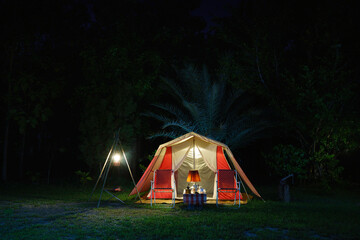 Camping tents at night, vintage cabin tents, antique oil lamps, retro chairs, and a group of camping tents with outdoor coffee-making facilities on wooden tables in a forest camping area.