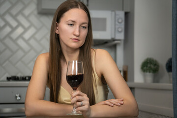 Young woman sitting alone at a kitchen table, holding a glass of red wine, looking pensive and reflecting on feelings of loneliness, sadness, and emotional turmoil