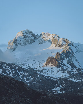 View of snow-capped peaks touched by the golden light of dawn, contrasting against the deep blue sky and rugged terrain, Cangas de Onis, Asturias, Spain.