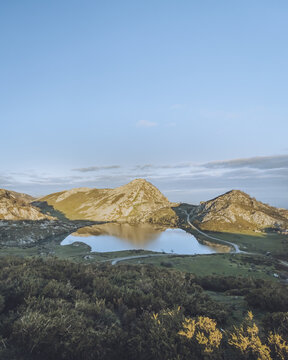 View of a serene lake mirroring the rugged peaks under a vast blue sky, framed by lush greenery in Lagos de Covadonga, Cangas de Onis, Asturias, Spain.