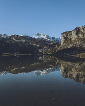 View of serene waters mirroring the snow-capped peaks and rugged cliffs under a clear blue sky, reflecting the stark beauty of nature, Cangas de Onis, Asturias, Spain.