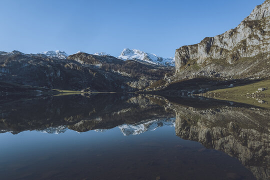 View of the tranquil waters mirroring the snow-capped mountains under a clear blue sky, reflecting the rugged cliffs in Lagos de Covadonga, Cangas de Onis, Asturias, Spain.