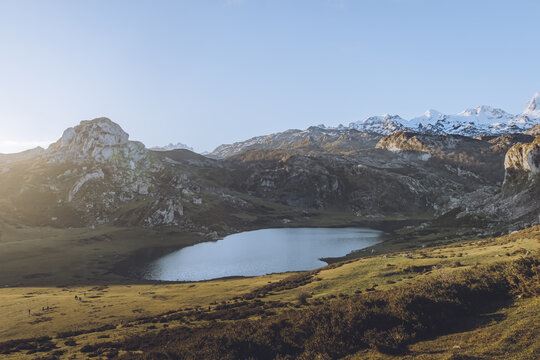 View of a serene lake nestled among rugged mountains, tinged with snow and sunlit slopes, creating a contrast of cool blues and warm earthy tones, Cangas de Onis, Asturias, Spain.