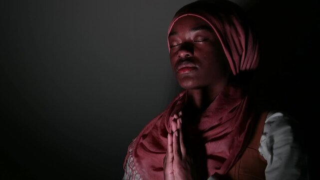 Cinematic low-key portrait of a young woman in a headscarf praying with closed hands, dark studio atmosphere highlighting spirituality, devotion, emotional depth, belief, and quiet inner reflection