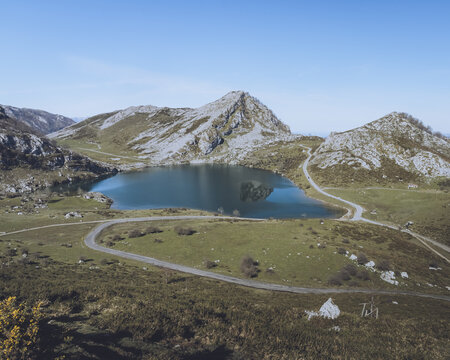 View of the serene Lago Enol mirroring rugged mountains under a clear sky, amidst winding roads and vibrant green meadows, Cangas de Onis, Asturias, Spain.