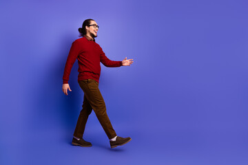 Young handsome man with glasses walks in a red sweater and brown pants smiling against a violet...
