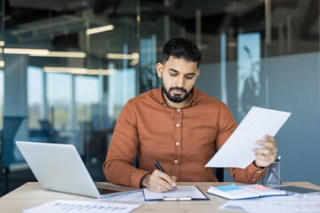 Fototapeta premium Businessman focused on reviewing and signing contract documents at a modern office desk with laptop and paperwork, conveying professional responsibility, negotiation and career-focused determination