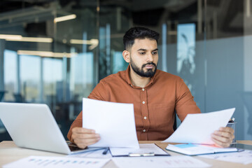 Obraz premium Young indian businessman intently analyzing financial charts and paperwork at a modern desk with laptop, preparing reports and planning strategy for corporate finance and projects