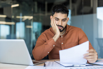 Stressed male employee experiencing frustration and overwhelm, holding document while feeling tired from heavy workload and endless paperwork in a modern office workplace