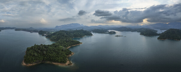Aerial view of lush green islands rise from the tranquil, dark waters under a dramatic, cloudy sky at Cheow Lan Lake, Bang Nai Si, Phang Nga, Thailand.