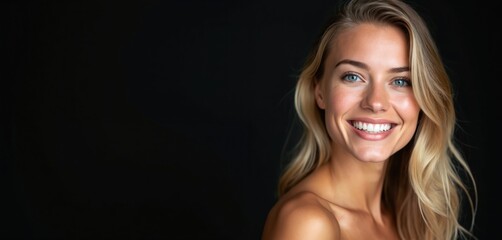 Fototapeta na wymiar Young happy blonde woman smiles into camera. She has radiant skin, bright blue eyes, and perfect white teeth. Natural beauty portrait on dark studio background. Female model showing healthy skin care.