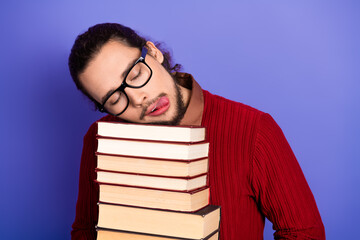 Young handsome student rests head on stack of book wearing glasses and sweater against purple...