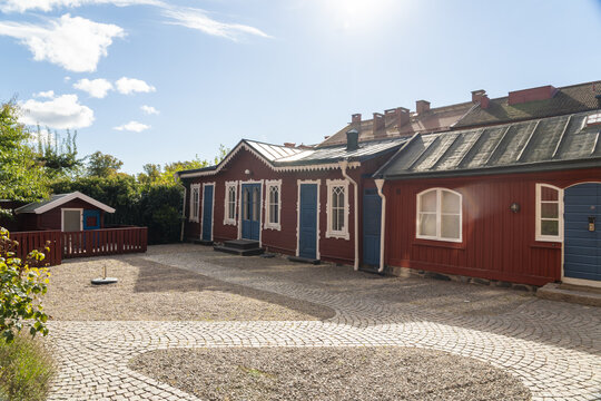 View of rustic red wooden buildings with ornate white trim and blue doors grace a gravel courtyard under a bright, blue sky, Karlskrona, Blekinge County, Sweden.