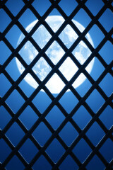 Abstract night composition featuring bright full moon seen through dark geometric grid structure against deep blue sky. Concepts of restriction, freedom, surveillance, protection, or design background