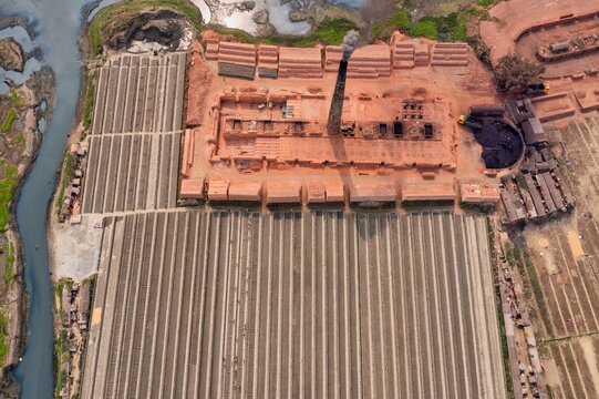Aerial view of brick factory with stacks of bricks, a tall chimney expelling smoke, and dark sludge contrasting against the landscape, Dhaka, Dhaka Division, Bangladesh.