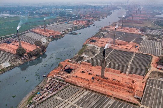 Aerial view of brick kilns lining the riverbanks, smoke billowing from chimneys under a hazy sky, Dhaka, Dhaka Division, Bangladesh.
