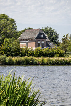 View of a quaint brick house with a dark roof peeks from behind a screen of lush green trees, reflected in the calm waters, Zaandam, North Holland, Netherlands.