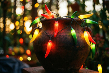 A festive string of chili pepper lights in red, yellow, and green, wrapped around a rustic clay pot with a warm, blurry bokeh background.