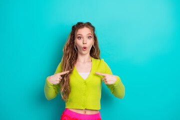 Young woman expressing surprise while pointing at herself against a turquoise background, showing...