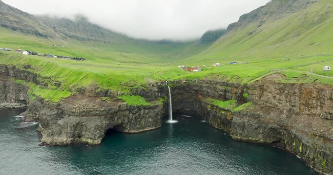 Aerial view of the stunning Mulafossur Waterfall cascading into the ocean from green cliffs, contrasting with the dark blue sea, Mulafossur, Faroes.