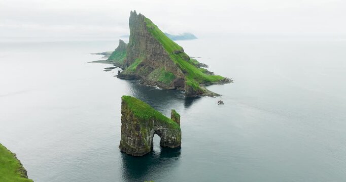 Aerial view of the dramatic Tindholmur islet with its lush green cliffs and jagged peaks surrounded by the vast, grey Atlantic ocean, Tindholmur, Faroes.