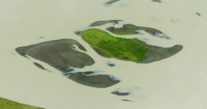 Aerial view of a glacial river shows a stark contrast between the pale water and the dark, rugged islands with green patches, Iceland.