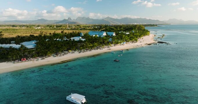 Aerial view of turquoise waters meet the white sand beach, fringed by green palms and white buildings under the distant mountains, Mauritius.