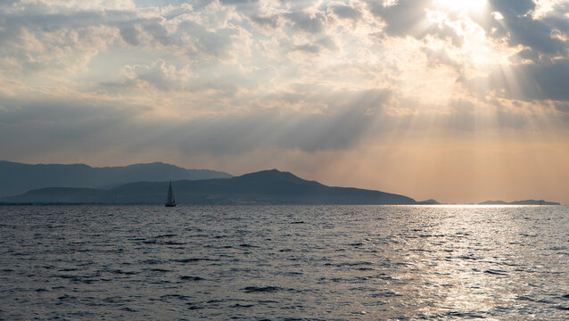 Sailboat on Calm SPeaceful seascape with a sailboat on calm water, distant mountains on the horizon and dramatic sun rays breaking through clouds. Golden sunset light reflecting on the sea surface. 