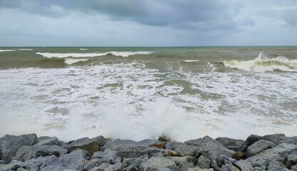 The beach is rough and rocky during the rainy season and the sky is cloudy. Ripples on the sea are the ocean surge. Clouds above waves on stone beach