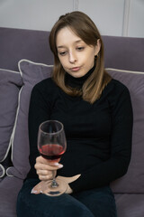 Young woman sitting on a sofa, holding a glass of red wine, wearing a melancholic expression while grappling with feelings of loneliness and emotional distress in her cozy home