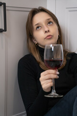Pensive young woman sitting on the kitchen floor, holding a glass of red wine and gazing upward, embodying feelings of loneliness and deep contemplation amidst her surroundings