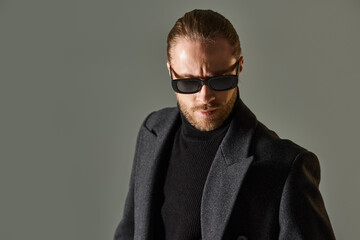 Obraz premium Handsome man in elegant coat and stylish sunglasses poses in grey studio setting