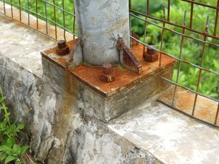 Extreme close-up of a heavily rusted metal base plate with large anchor bolts secured to a weathered concrete foundation with green growth.