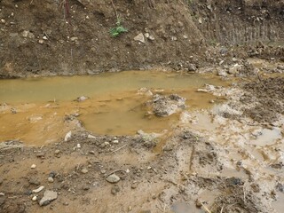 A very close-up detail of a muddy ditch filled with brown, opaque water and large clumps of churned earth, rocks, and visible tire tracks.