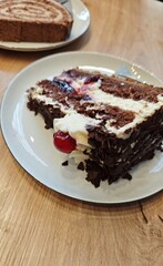 Chocolate cake slice with cream layers, cherry filling and chocolate shavings served on a plate on a wooden table. Concept of dessert indulgence, sweet treat enjoyment and cozy cafe moment