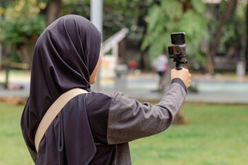 A young Indonesian Muslim woman recording a vlog video using an action camera in a city park
