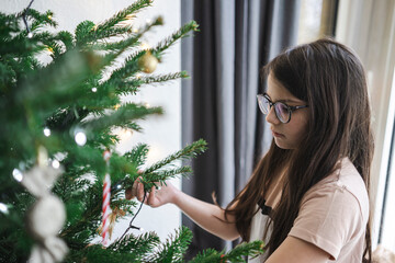 Teenage girl in glasses and T-shirt seriously decorating Christmas tree at home, focused holiday moment