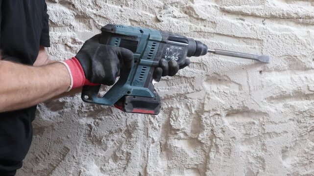 Close-up of worker using electric hammer drill to remove old plaster from concrete interior wall. Renovation and demolition process with rough wall texture, dust and damaged surface. Concept of home i