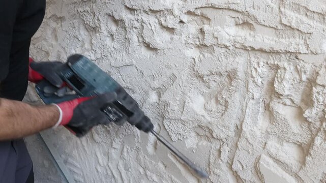 Close-up of worker hands using electric hammer drill to remove old plaster from interior concrete wall. Renovation and demolition process with rough texture, dust and damaged surface. Concept of home 
