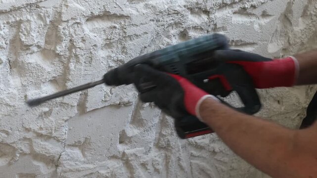 Close-up of worker hands using electric hammer drill to remove old plaster from interior concrete wall. Renovation and demolition process with rough texture, dust and damaged surface. Concept of home 