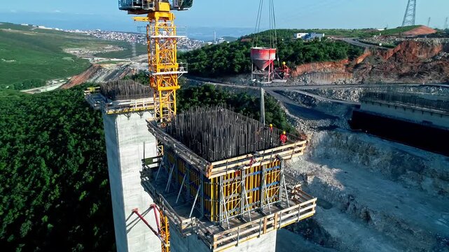 Drone aerial view of reinforced concrete bridge pier head construction with formwork, rebar and tower crane. Major highway viaduct infrastructure project in mountainous terrain.