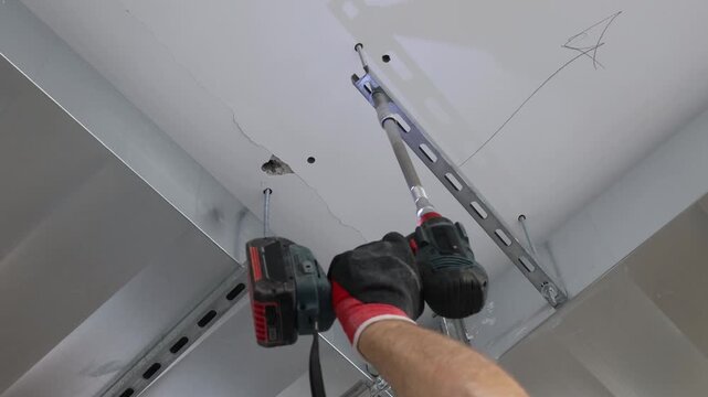 Close-up of construction worker installing metal ceiling frame using cordless drill. Professional drywall ceiling installation, interior renovation process, suspended ceiling system, building construc