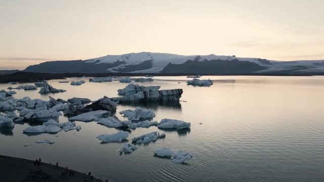 Golden hour aerial flight reflecting warm sunset light on blue ice chunks at J&ouml;kuls&aacute;rl&oacute;n lagoon, Iceland.